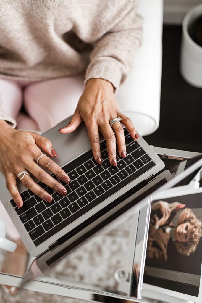 Black woman typing on computer with coffee sitting next to her wearing a cream sweater and light pink pants