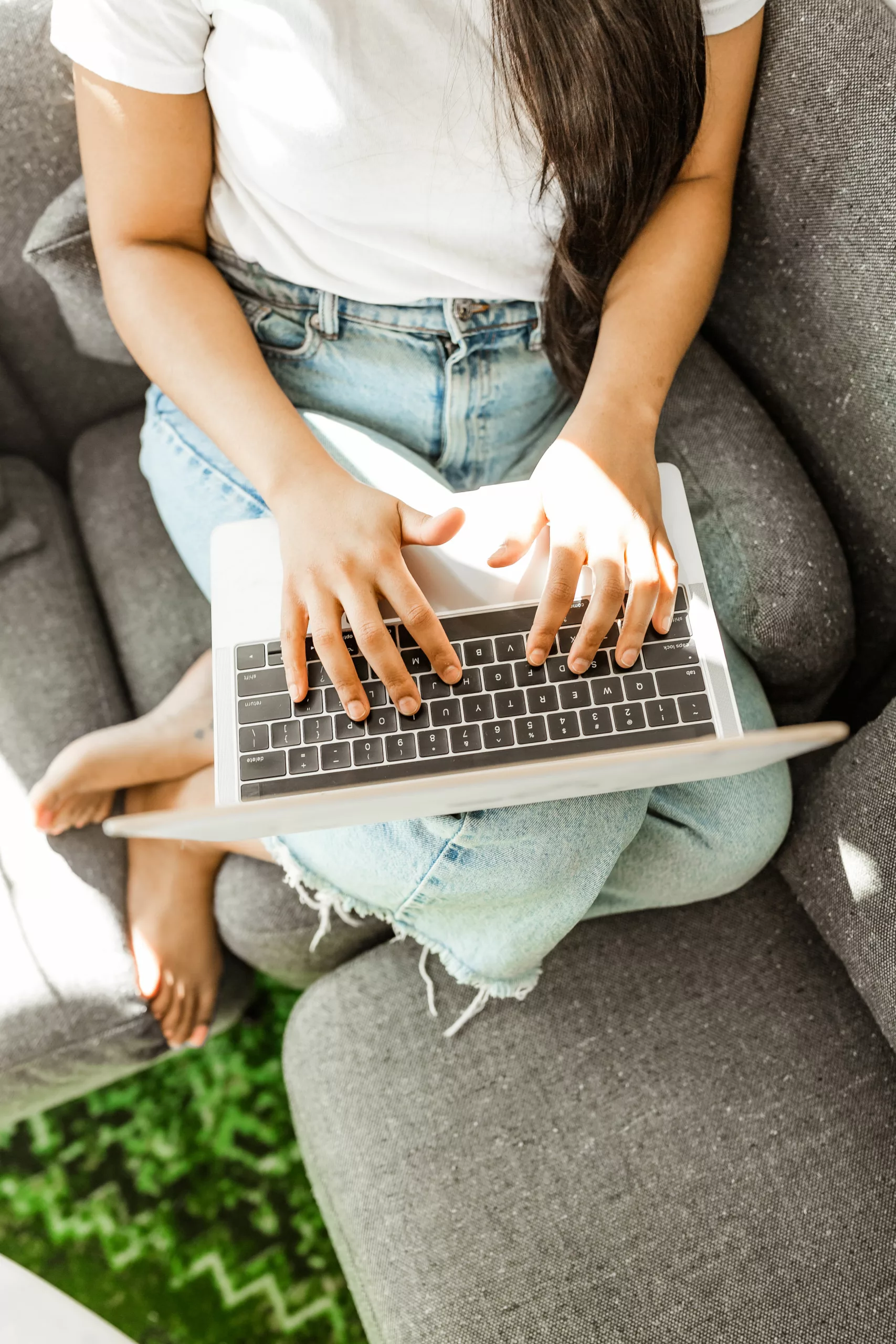 Woman on the couch with blue jeans and a white shirt using her macbook to type.