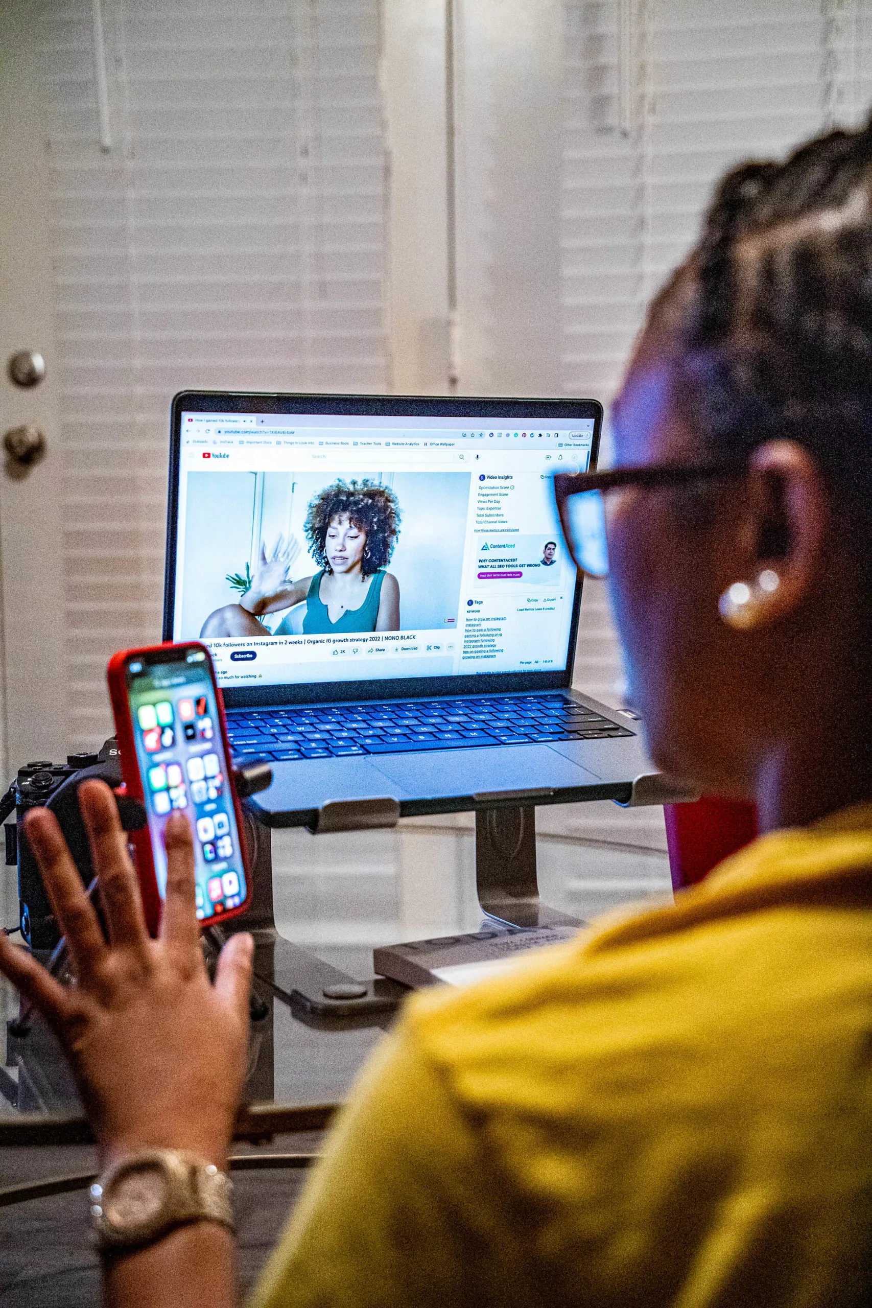 Black woman in yellow shirt using cellphone and laptop simultaneously.