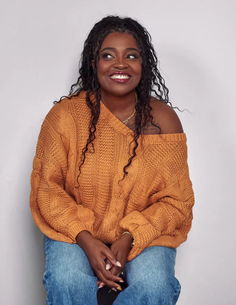 Black woman sitting on stool in a burnt orange sweater looking to the side smiling
