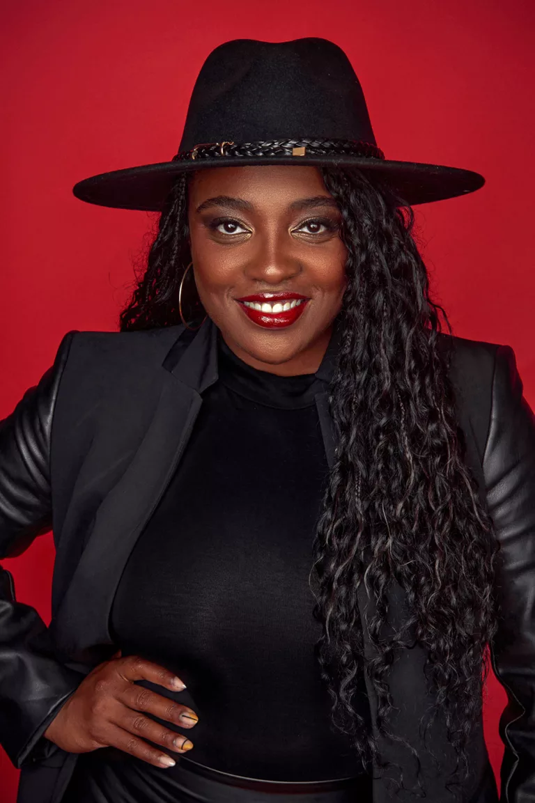 Black woman dressed in all black with a fidora against a red wall.