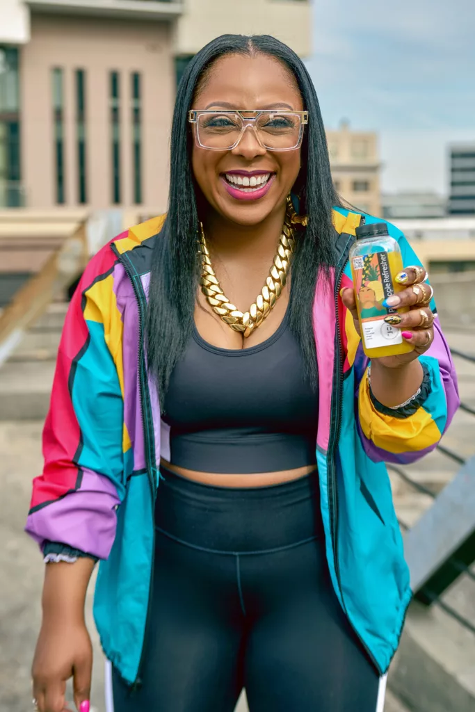 woman in black athletic wear holding a bottle of juice wearing gold jewelry and a colorful jacket..