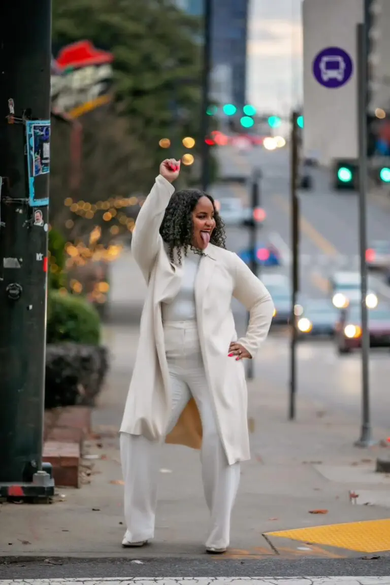 Woman standing on the sidewalk in the city with a cream colored trench coast, cream turtle neck and cream pants. Laughing with her tongue out of her mouth.