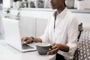 Woman sitting at table wearing a white button up shirt using laptop and drinking coffee