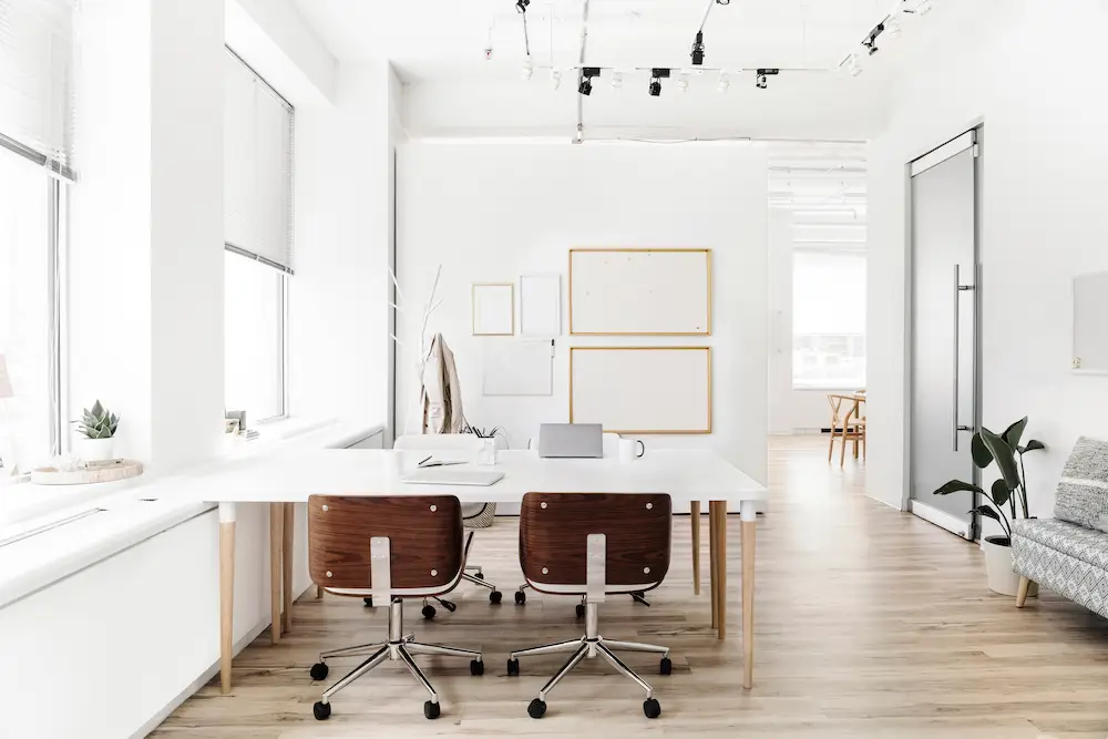 Minimalist office with white walls. white desk and a white chair all sitting by aa wall with three windows.